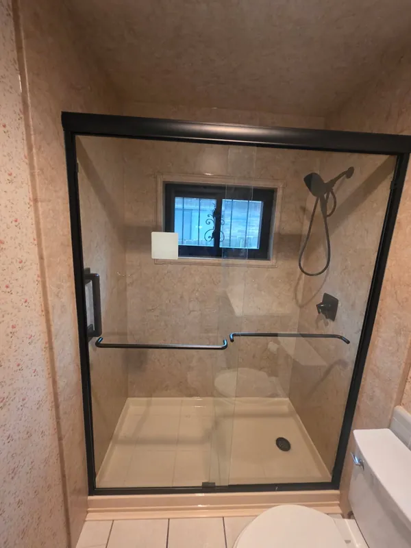 Beige marble-pattern walk-in shower with black-framed bypass glass doors, oil-rubbed bronze handheld and dual grab bars over a low-profile pan