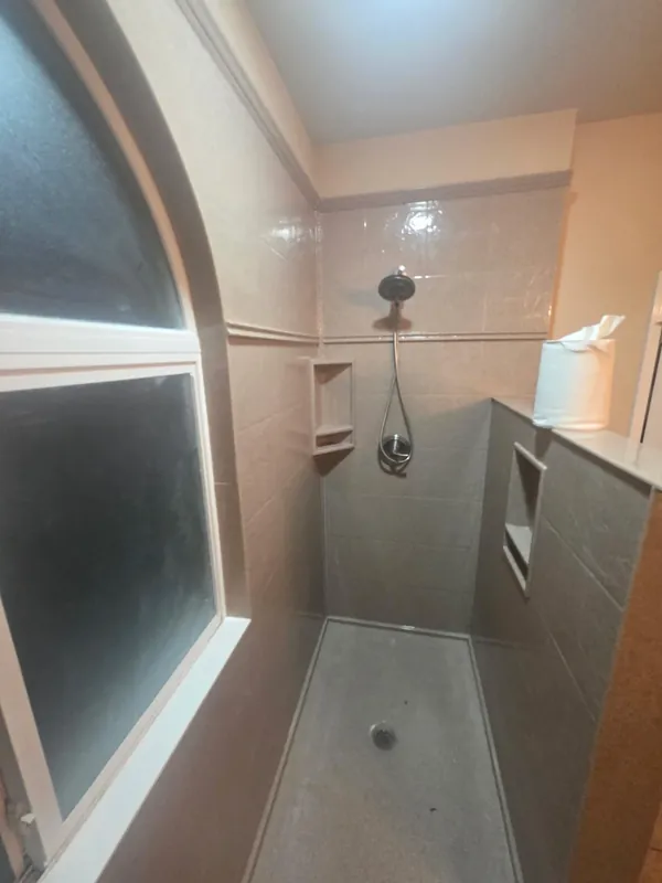 Curbless walk-in shower with two-tone tile, recessed soap niche and arched glass-block window — full bathroom conversion by our crew