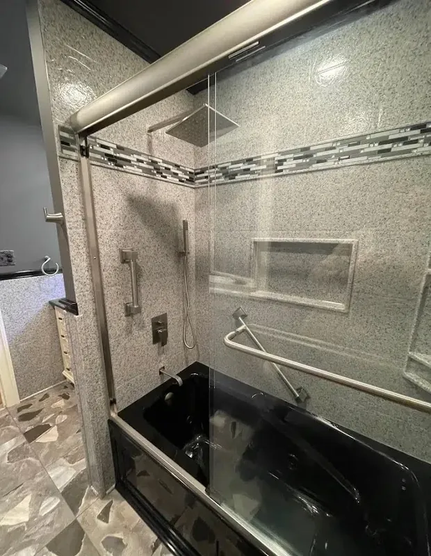 Black soaking tub with granite-pattern surround, glass mosaic accent band, frameless glass shield and brushed-nickel grab bars