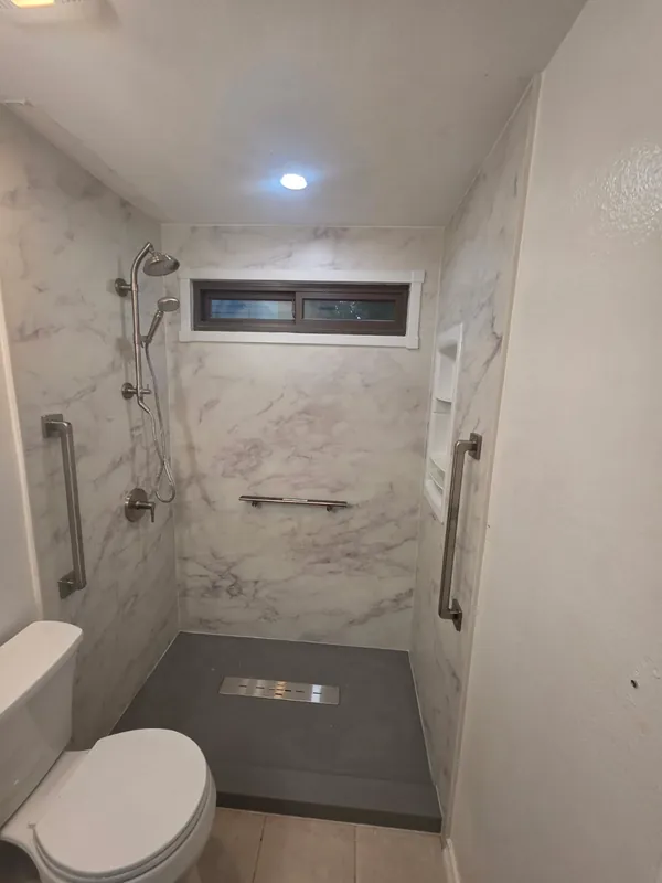 Curbless white marble-pattern walk-in shower with linear drain, brushed-nickel rain shower, dual grab bars and recessed corner shelves for safe aging-in-place bathing